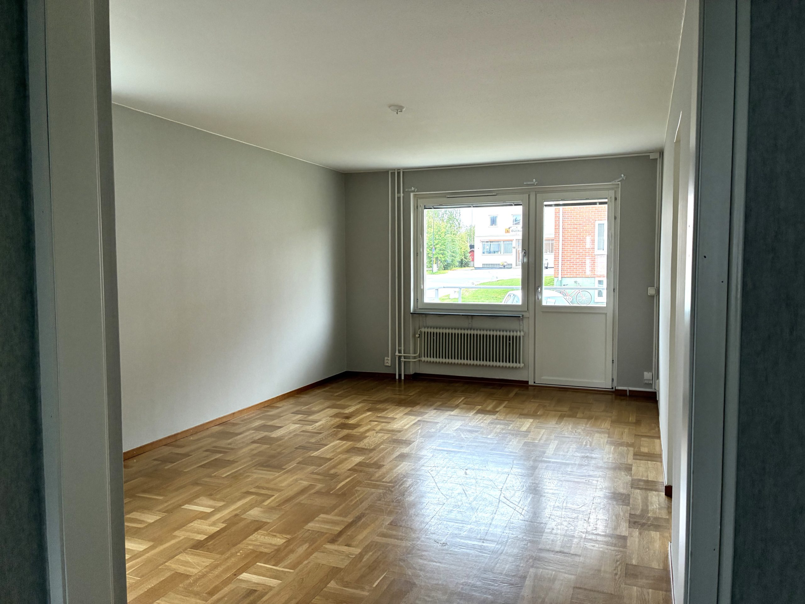 Vardagsrum med ljusa väggar och träfärgat golv. På väggen längst bort i bild finns ett fönster och en balkongdörr.