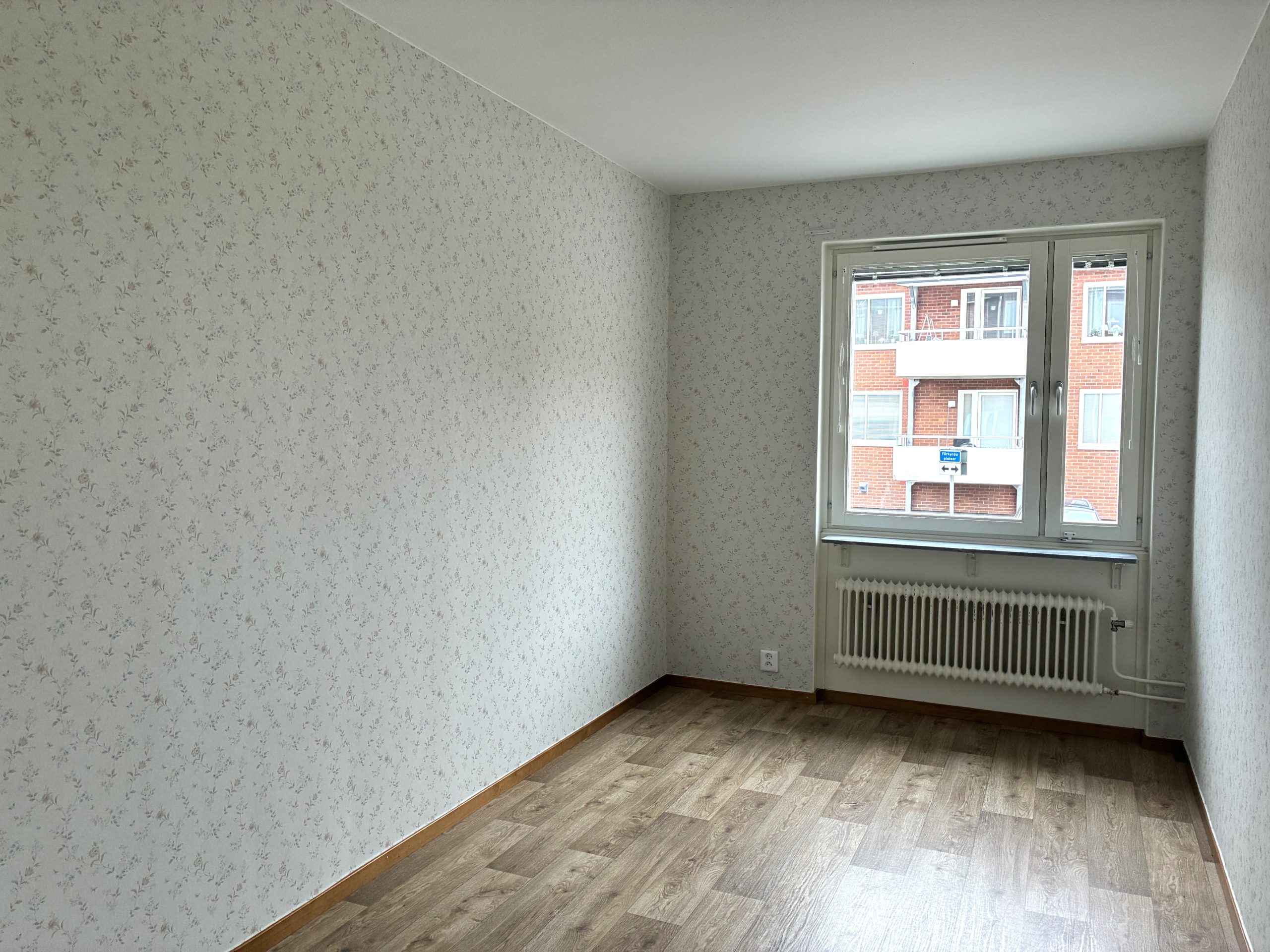 Sovrum med blommiga tapeter och ett träfärgat golv. På väggen längst bort finns ett fönster.