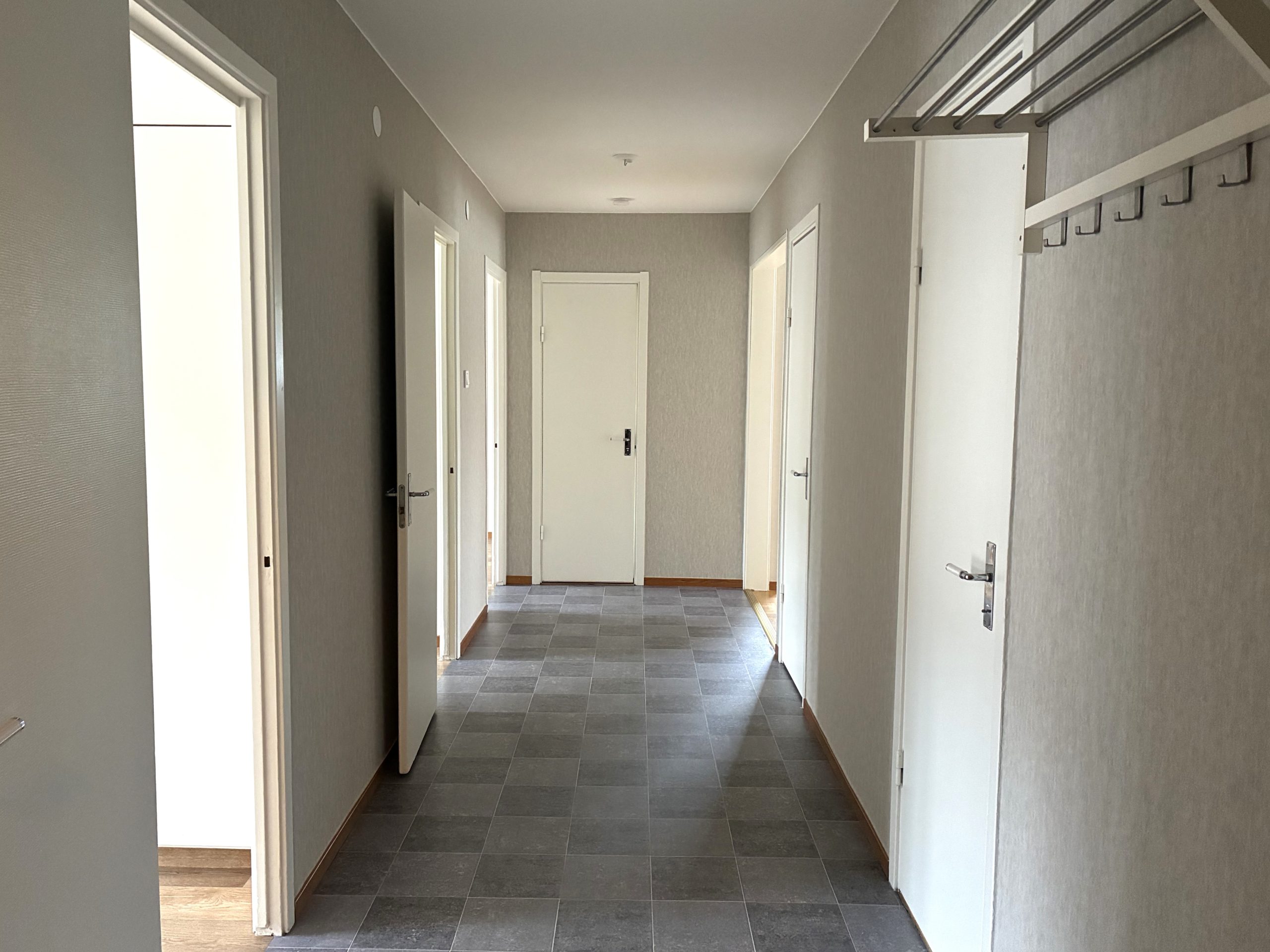 Hall med grå väggar och grå golmatta. Flertalet vita innerdörrar leder till lägenhetens olika rum.