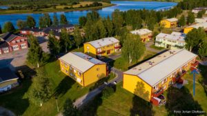 Flygfoto över ett mindre bostadsområde med gula hyreshus som omgärdas av grönområden och träd. i bakgrunden syns andra byggnader och ett vattendrag. En blå pil pekar mot en specifik lägenhet i det främre hyreshuset.