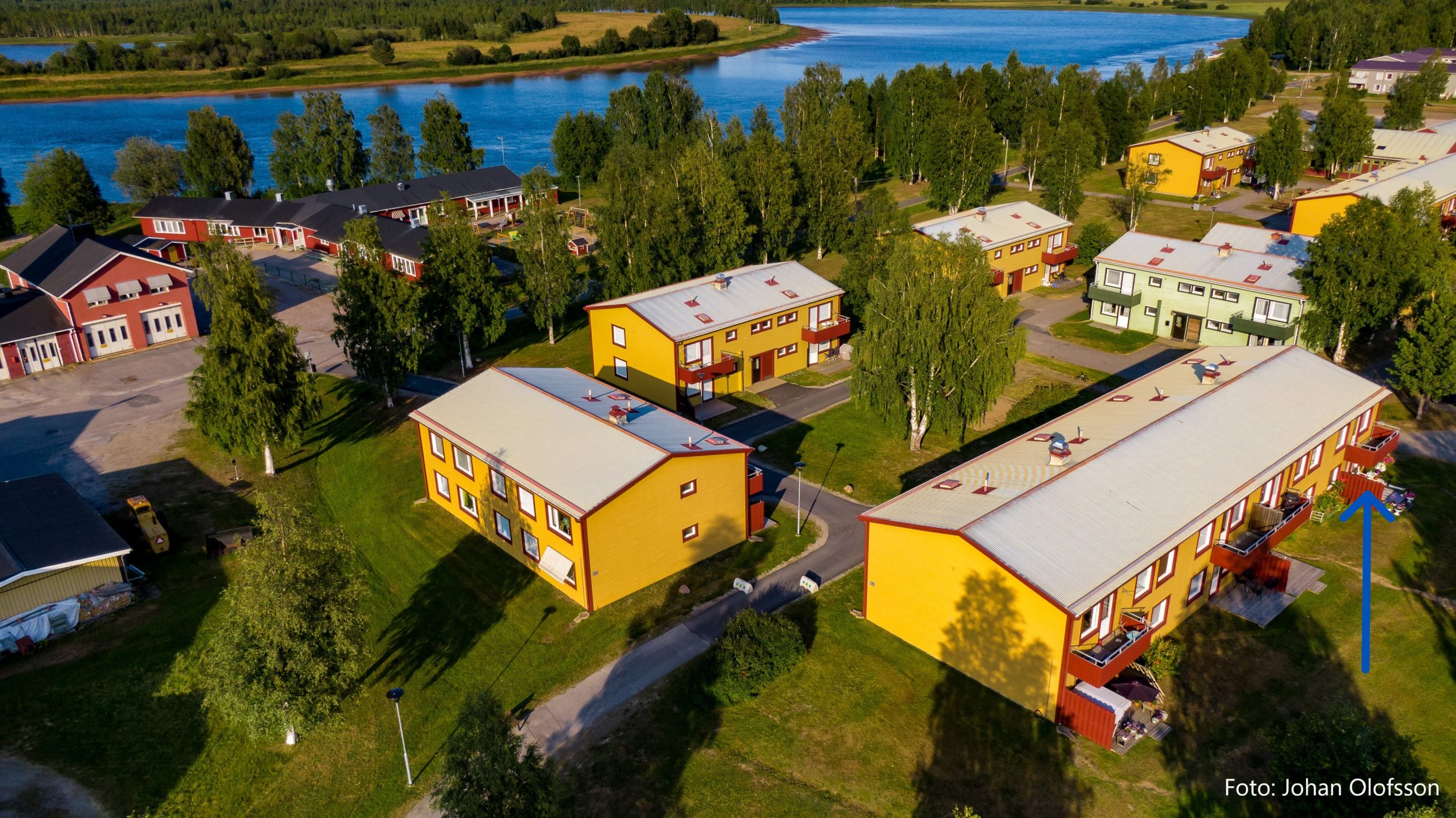 Flygfoto över ett mindre bostadsområde med gula hyreshus som omgärdas av grönområden och träd. i bakgrunden syns andra byggnader och ett vattendrag. En blå pil pekar mot en specifik lägenhet i det främre hyreshuset.
