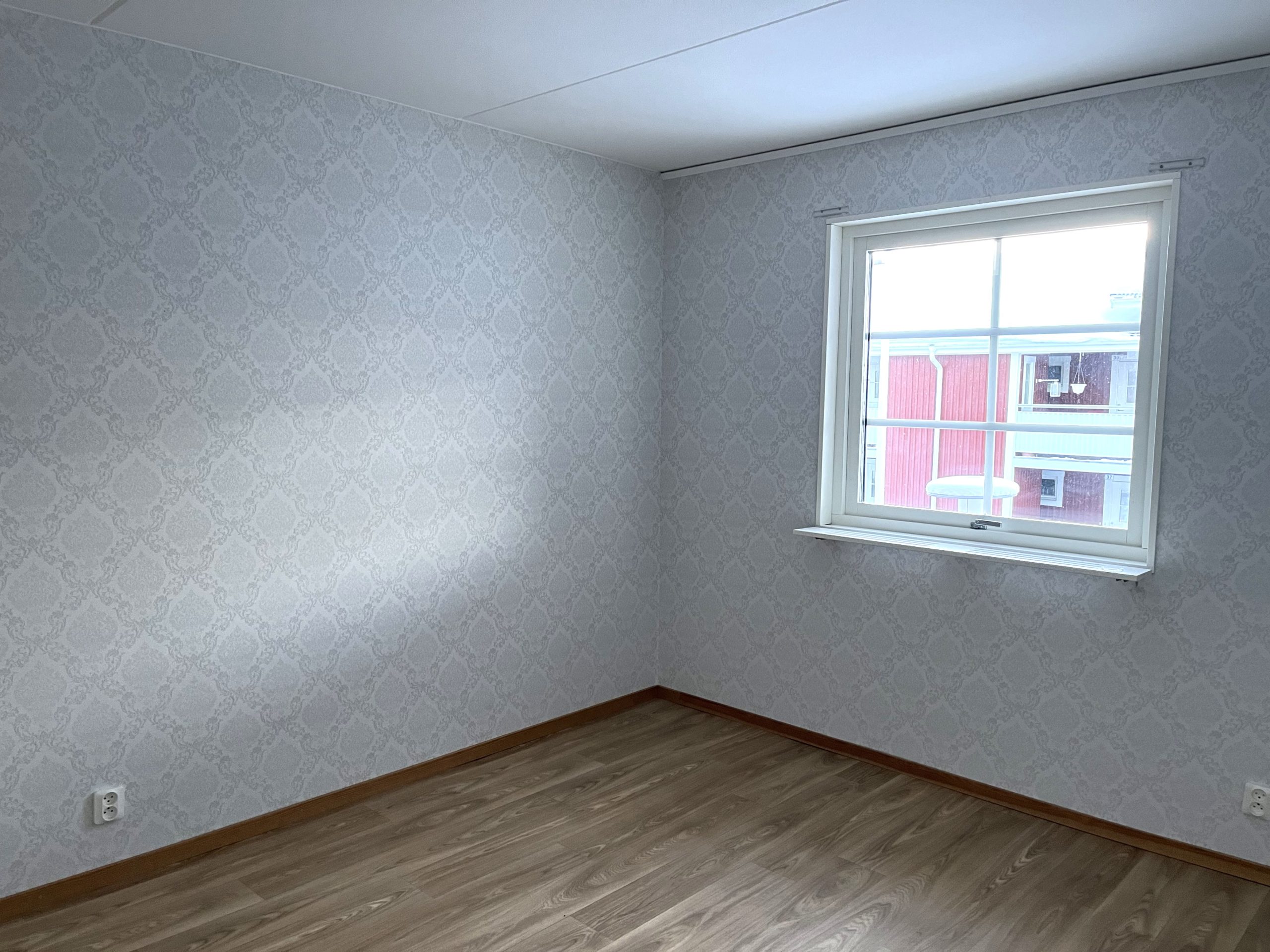 Sovrum med ljusa tapeter och ett träfärgat golv. På den bortre väggen finns ett fönster.