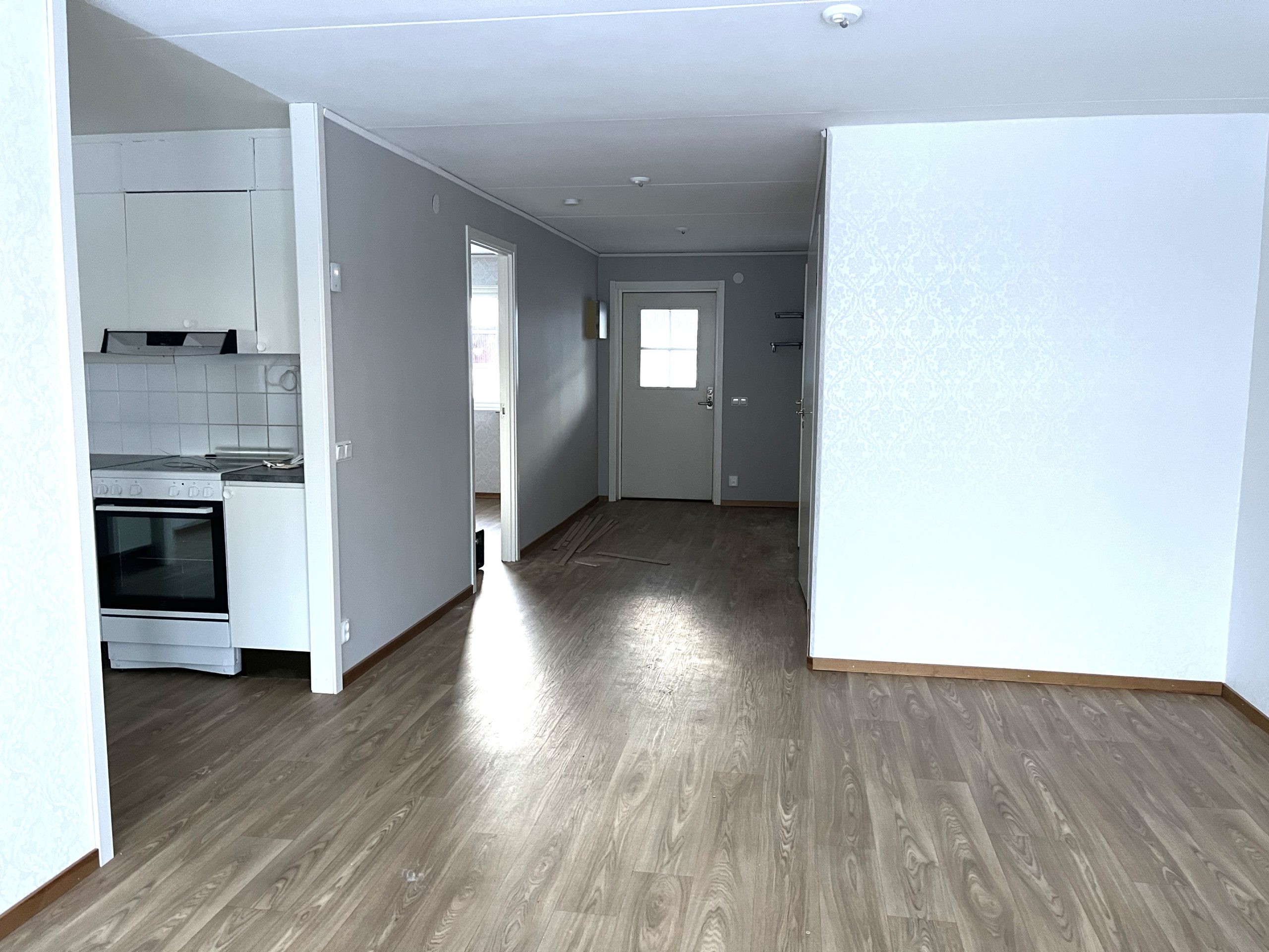 Foto från ett vardagsrum med öppen planslösning där man skymtar kök och hallen som leder bort till ytterdörren. Ljusa tapeter och ett träfärgat golv