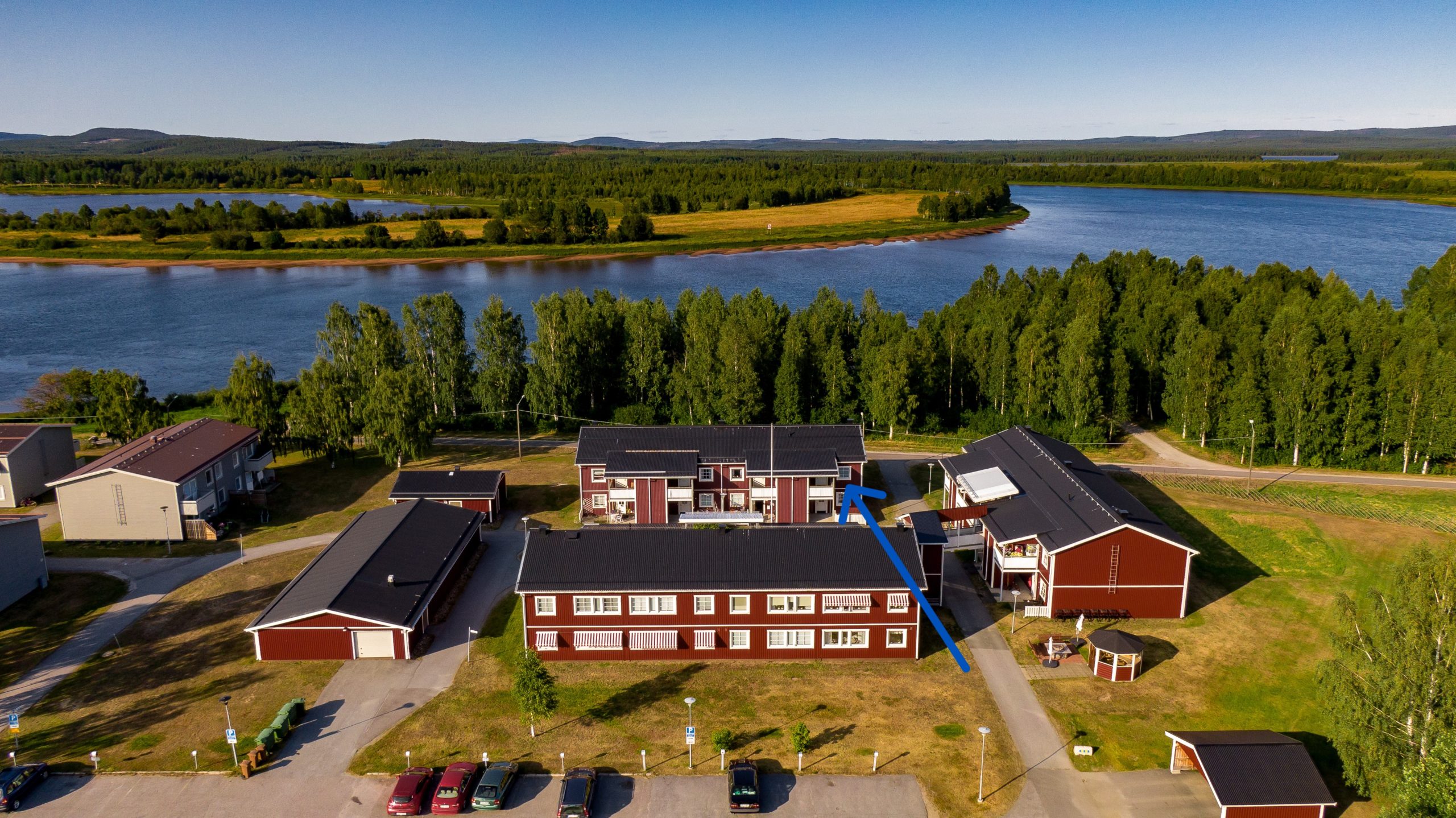 Flygfoto över ett kluster av röda byggnader nära en älv, omgiven av träd och gräsytor. En pil markerar en särskild lägenhet i en av fastigheterna. Landskapet är platt med avlägsna kullar som erbjuder fridfulla vyer.