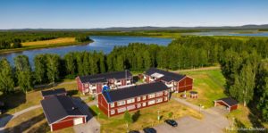 Flygfoto över ett kluster av röda byggnader nära en älv, omgiven av träd och gräsytor. En pil markerar en särskild lägenhet i en av fastigheterna. Landskapet är platt med avlägsna kullar som erbjuder fridfulla vyer.