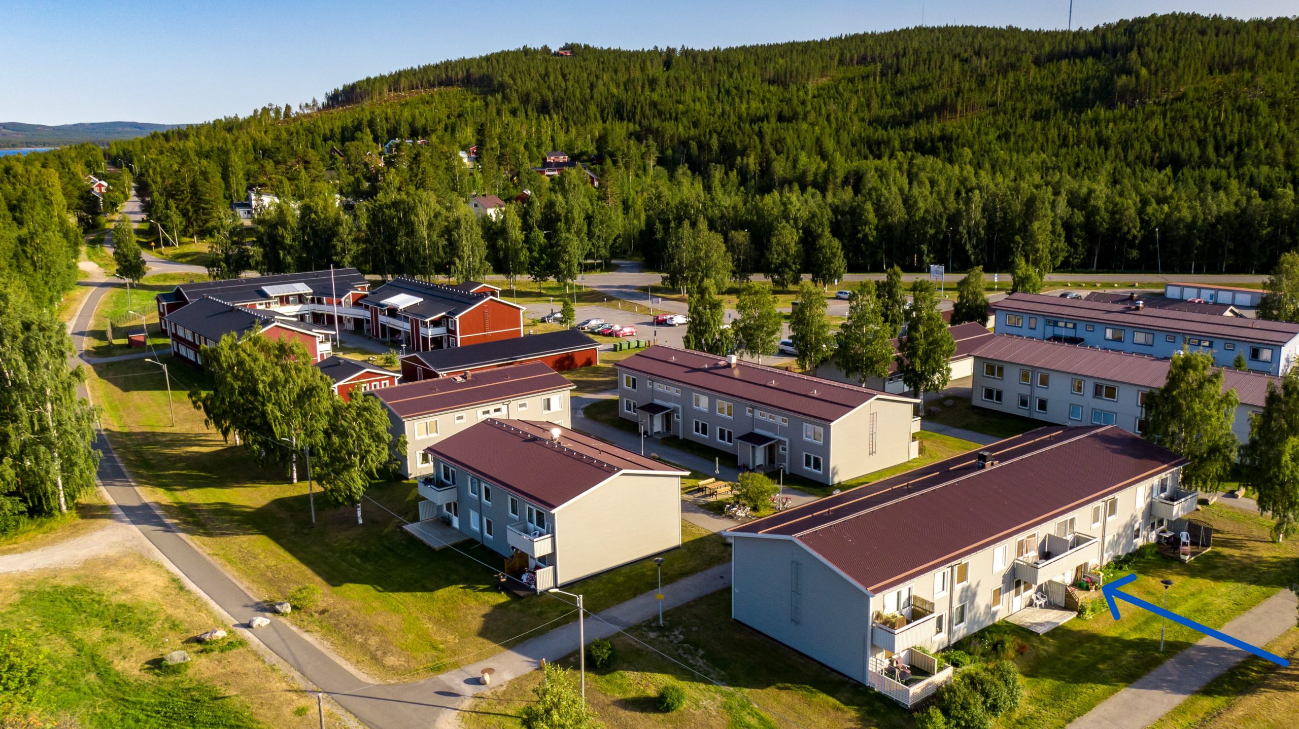 Flygfoto över ett bostadsområde med flera moderna hyreshus omgivna av grönskande träd och gräsytor. Ett nätverk av stigar är synligt och tät skog kan ses i bakgrunden under en klarblå himmel.