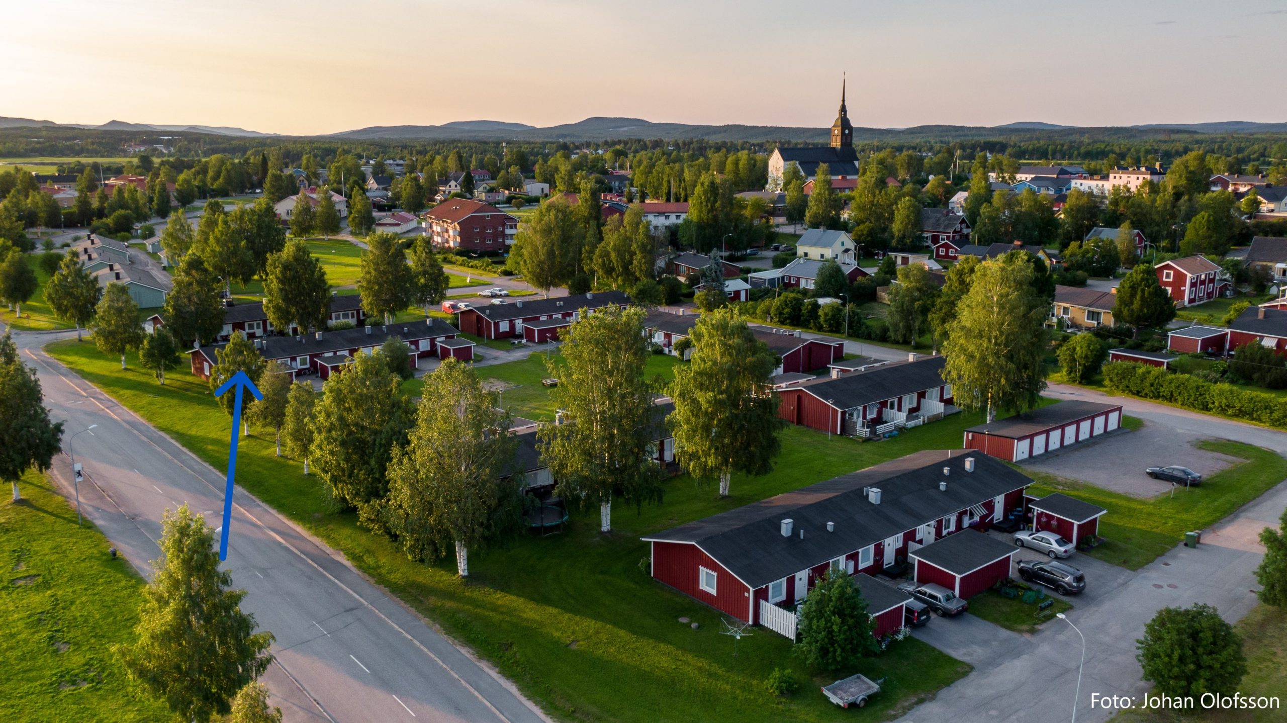 Flygfoto över ett bostadsområde med enplanshus. Husen står på ett grönområde med träd och i bakgrunden skymtar ett mindre samhälle med en kyrka i blickfånget. En bilväg skymtar till vänster i bild och i fjärran finns berg. En blå pil pekar mot en specifik lägenhet.