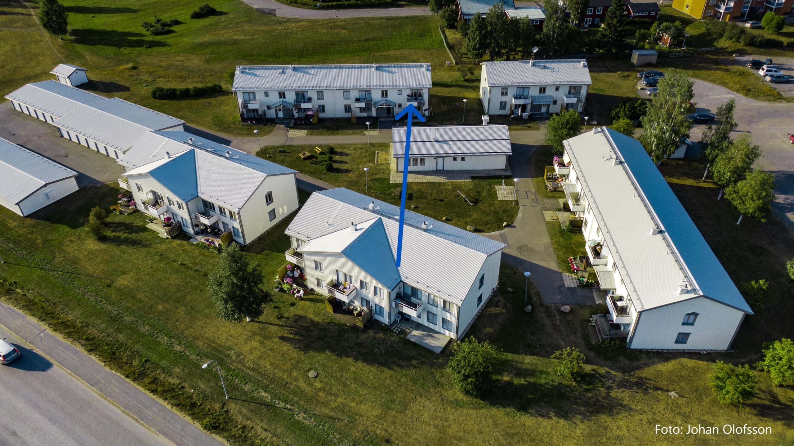 Flygfoto över ett bostadsområde med flera vita byggnader omgivna av grönt gräs och träd. En blå pil pekar mot en av byggnaderna.