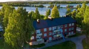 Ett brunt lägenhetshus med svart tak med fönster och flertalet balkonger. I bakgrunden skymtar en älv, träd och kullar. En blå pil pekar mot en specifik lägenhet i fastigheten.
