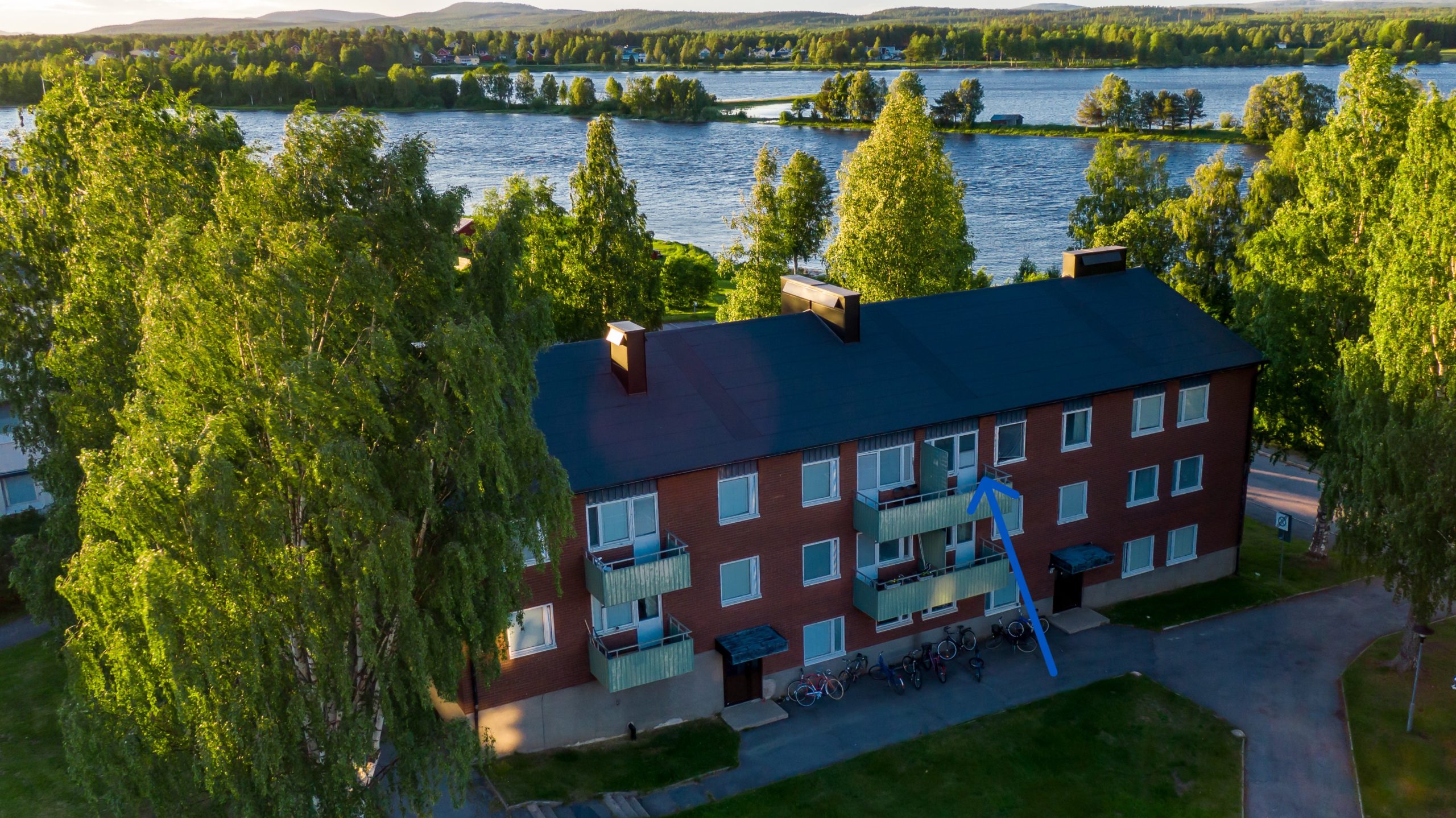 Ett brunt lägenhetshus med svart tak med fönster och flertalet balkonger. I bakgrunden skymtar en älv, träd och kullar. En blå pil pekar mot en specifik lägenhet i fastigheten.