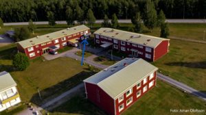 Flygfoto över tre röda bostadshus, med ljusa tak omgivna av grönskande gräsmattor och träd. En bil står parkerad i närheten, medan en trädkantad väg sträcker sig i bakgrunden.