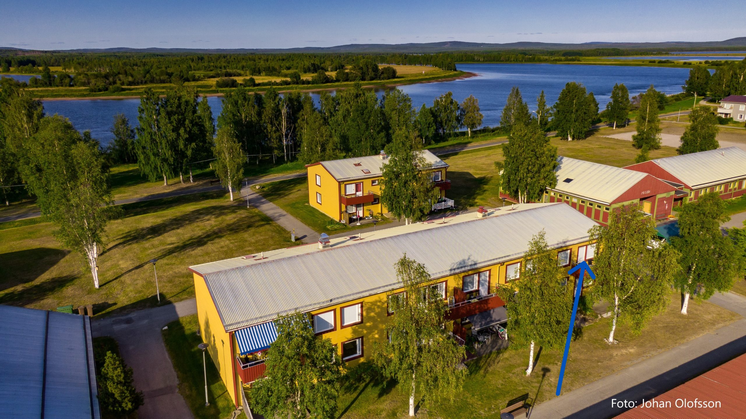 Flygfoto över ett bostadsområde med färgglada byggnader omgivna av träd och grönska. En älv rinner i bakgrunden under en klarblå himmel. En blå pil pekar på en specifik lägenhet i en gul byggnad.