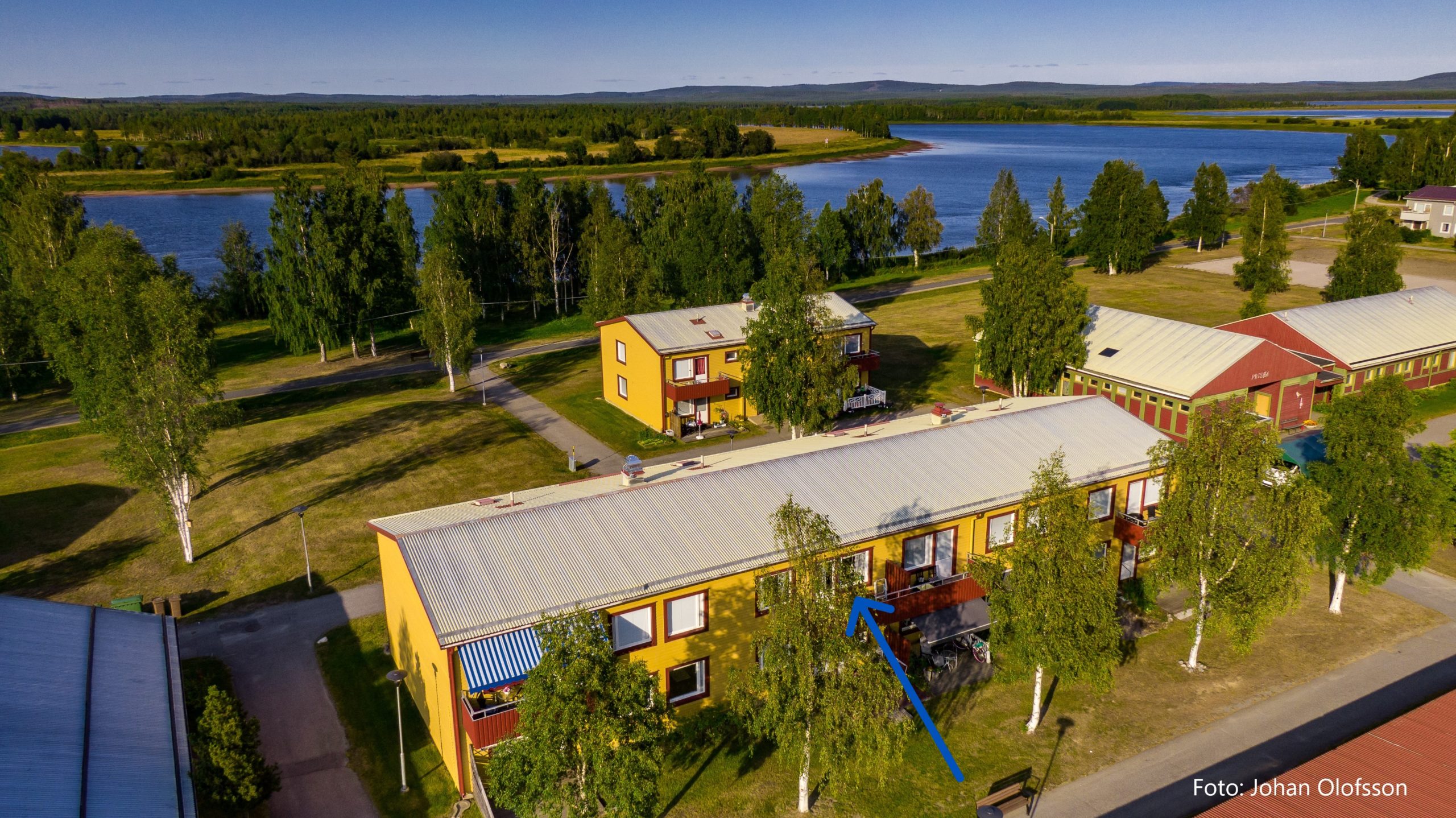 Flygfoto över ett bostadsområde med färgglada byggnader omgivna av träd och grönska. En älv rinner i bakgrunden under en klarblå himmel. En blå pil pekar på en specifik lägenhet i en gul byggnad.