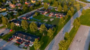 Flygfoto över ett bostadsområde med enplanshus. Husen står på ett grönområde med träd. En bilväg skymtar till höger i bild. En blå pil pekar mot en specifik lägenhet.