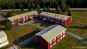 Flygfoto över tre röda byggnader nära en väg, omgiven av träd och gräsytor. En pil markerar en särskild lägenhet i en av fastigheterna.