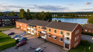 Flygfoto över ett tvåvåningshus av tegel. Parkeringsplatser och människor syns utanför. En blå pil pekar på en specifik port. Träd och en älv syns i bakgrunden under en delvis molnig himmel.