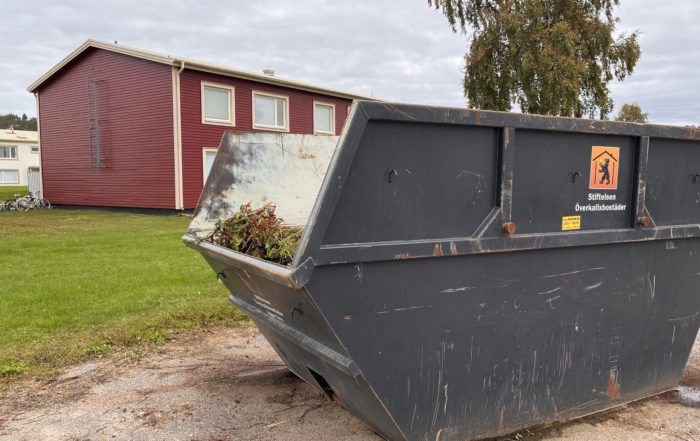 Bilden visar en container märkt med Stiftelsen Överkalixbostäder. I bakgrunden syns ett rött lägenhetshus.